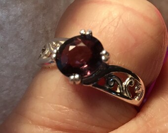 May include: A silver ring with an intricate design featuring a dark purple gemstone.