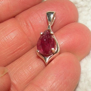 May include: A pear-shaped pink gemstone pendant set in a silver setting. The pendant is on a white surface.