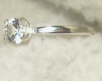 May include: A silver ring with a clear, round gemstone. The gemstone is set in a four-prong setting, and the band is smooth and polished. The ring is likely made of sterling silver or a similar metal.