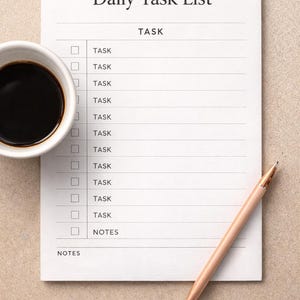 Puede incluir: Un bloc de notas blanco de Lista de tareas diarias con el título en la parte superior y la palabra "TAREA" junto a las casillas de verificación. Una taza de café negro y un bolígrafo están al lado.