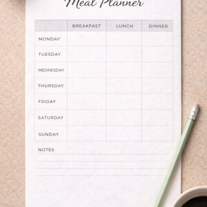 Puede incluir: Un planificador de comidas blanco con el título "Meal Planner". El planificador incluye secciones para cada día y horarios de comidas. Un lápiz verde claro y una taza de café están al lado.