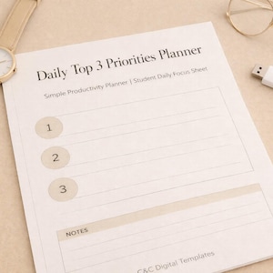 May include: A beige-toned desk scene featuring a "Daily Top 3 Priorities Planner" with numbered sections. A gold-toned watch, glasses, a USB drive, and a pencil are arranged around the planner, suggesting a focus on productivity and organization.