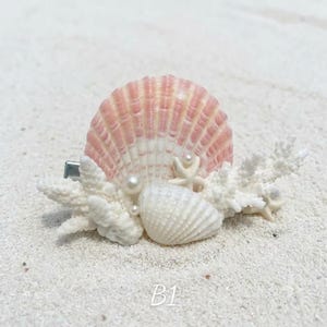 May include: A hair clip featuring a large, fan-shaped pink and white scallop shell, a smaller white seashell, and white coral-like branches. The accessory is decorated with small, round white pearls and a tiny starfish, all attached to a metal clip.