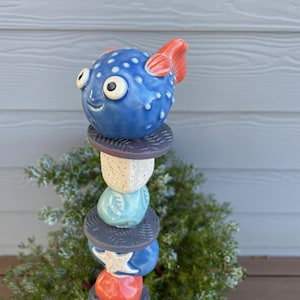 May include: A whimsical garden stake featuring a blue ceramic pufferfish with orange fins atop a stack of colorful ceramic shapes. The stake includes a white star, a red sphere, and various textured elements, perfect for outdoor decor.