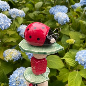 May include: A vertical garden stake featuring a ceramic ladybug at the top, followed by a stack of alternating green leaf-shaped discs, speckled beige spheres, and a red cube. A second, smaller ladybug is positioned lower on the stack.