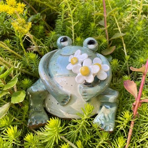 May include: A ceramic frog figurine with a glossy light blue and green speckled glaze, featuring two large eyes and a wide smile. The frog is holding a small bouquet of three white flowers with yellow centers in its front paws.
