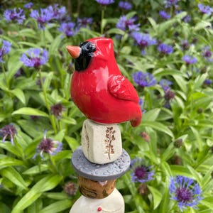 May include: A vibrant red ceramic cardinal bird figurine perched atop a stack of decorative, textured stones. The bird has a black mask and beak. The stones are various colors and shapes, with floral and dot patterns. The background features green foliage and purple flowers.