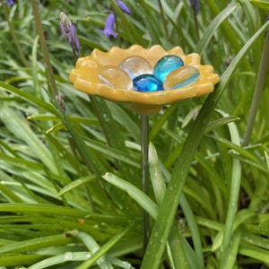 May include: A yellow, flower-shaped garden stake with a metal stem. The flower holds clear and blue glass stones. The stake is surrounded by green grass and purple flowers.