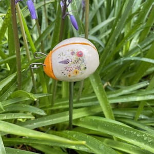 May include: A garden stake featuring a white, oval-shaped design with a floral print. The stake has orange accents and metal antennae. It is set amidst green foliage and purple flowers, creating a natural garden theme.