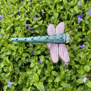 May include: A handcrafted dragonfly sculpture with a teal body and lavender wings. The dragonfly has small, silver-colored accents along its body and is set against a backdrop of green foliage with small purple flowers.