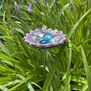 May include: A purple ceramic flower-shaped garden ornament with a metal stem, holding clear and blue glass beads. The ornament is surrounded by green grass and bluebell flowers, creating a natural garden aesthetic.