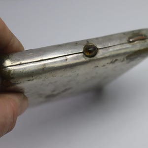 May include: A silver-colored, rectangular case with a tarnished finish. The case has a small, round, amber-colored button on the front and a hinged closure. The item is held by a hand.