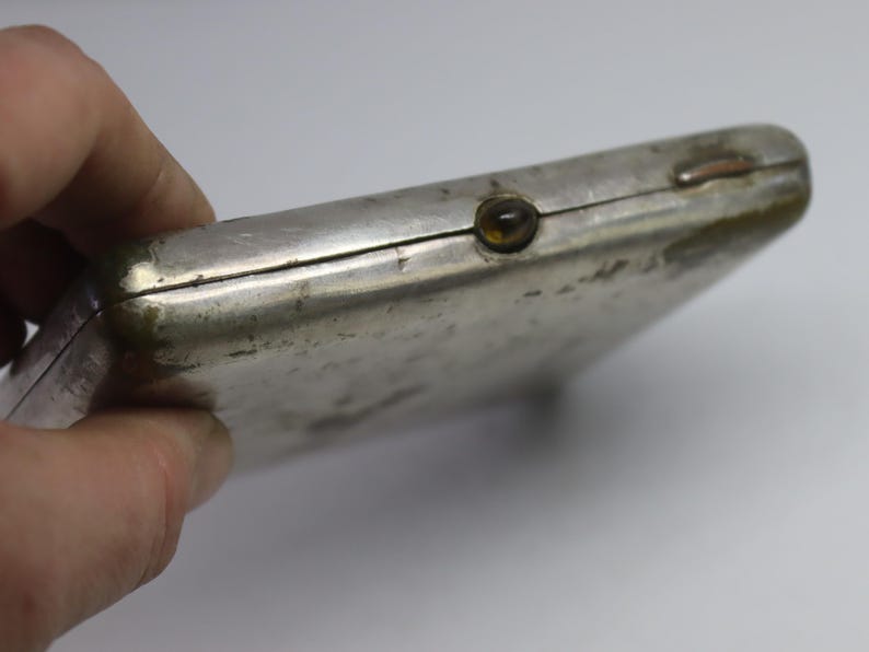 May include: A silver-colored, rectangular case with a tarnished finish. The case has a small, round, amber-colored button on the front and a hinged closure. The item is held by a hand.