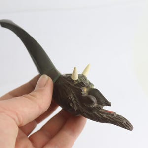 May include: A carved tobacco pipe with a demonic face design, featuring horns and a curved stem. The pipe is mainly brown with off-white horns and a dark green stem. The face has detailed features.