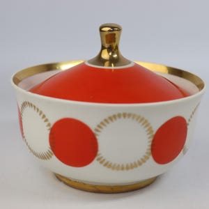 May include: A white ceramic sugar bowl with a red lid and gold detailing. The bowl has a pattern of red circles and gold-lined white circles. The lid has a gold knob. The bowl sits on a gold base.