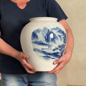 Pode incluir: Um vaso de cerâmica branco, grande e redondo, com um acabamento mate texturizado. A parte frontal apresenta um painel oval liso decorado com uma pintura de paisagem detalhada em azul e branco, retratando montanhas, árvores e casas.