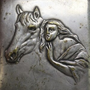 May include: Silver-toned metal case with a detailed relief of a horse's head and a woman. The horse's features are highlighted with gold accents, as is the woman's hair and clothing. The woman's expression is one of concern, and she is touching the horse.