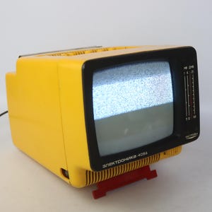 Puede incluir: Un televisor portátil de color amarillo brillante con un marco de pantalla negro que muestra estática. El dispositivo cuenta con deslizadores verticales de sintonización de canales a la derecha y el texto cirílico "Электроника-409Д" en el frente. Descansa sobre un soporte rojo.