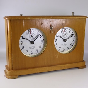 May include: A vintage wooden chess clock with two round clock faces. The clock has a light brown finish and features black hands and white clock faces with black numerals. The clock is marked "Moscow" in Russian.