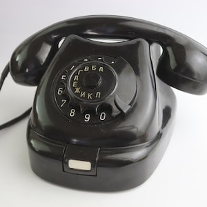 Puede incluir: Un teléfono negro vintage con disco giratorio y acabado brillante. El teléfono tiene un dial circular con números y letras cirílicas, un auricular y una base con un pequeño botón rectangular. El cable del teléfono es visible.