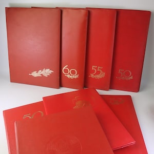 May include: A collection of red, leather-bound books. Some books have gold lettering and designs, including the numbers 50, 55, and 60. The books are arranged in a neat stack, with some lying flat and others standing upright.