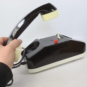 May include: A vintage black and cream-colored telephone with a handset. The phone has a red button and a white switch. The handset is held up, showing the cream-colored earpiece and mouthpiece.