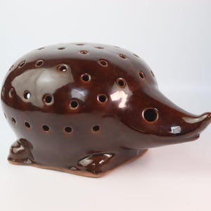 Può includere: Un portacandele a forma di riccio in ceramica marrone. Il pezzo decorativo ha una finitura lucida e numerosi piccoli fori sulla schiena. Il riccio ha un muso appuntito e zampette, creando un design stravagante.