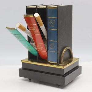 May include: A vintage cigarette dispenser designed as a miniature bookshelf with three book-shaped compartments. The unit features a black base, a brass trim, and three hinged book spines in green, red, and blue with gold Cyrillic text.