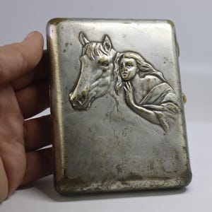 May include: A rectangular, silver-toned cigarette case with a detailed relief of a horse's head and a woman. The woman is draped in flowing fabric, and the case has a tarnished appearance.