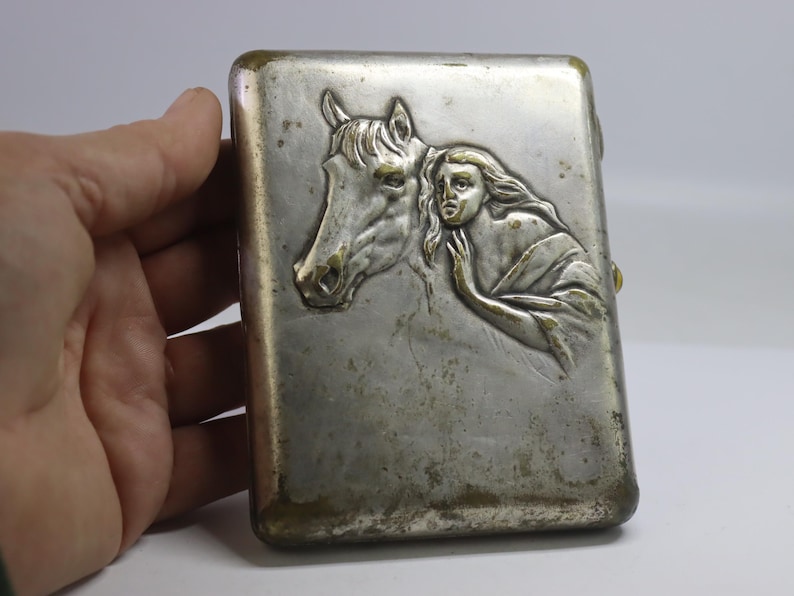 May include: A rectangular, silver-toned cigarette case with a detailed relief of a horse's head and a woman. The woman is draped in flowing fabric, and the case has a tarnished appearance.