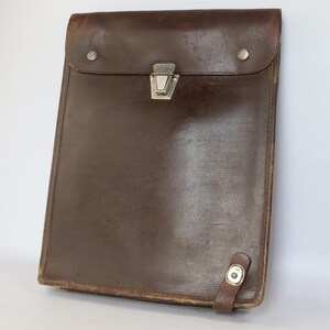 May include: A dark brown leather document case with a rectangular shape, featuring a fold-over flap secured by a silver-toned metal latch. The case includes two silver-toned rivets on the flap and a small leather strap with a metal snap closure.
