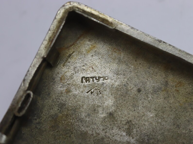 May include: Close-up of a rectangular, silver-toned metal object. The interior surface has a textured appearance with the word "PATYHO" and a symbol with the number "13" etched into the metal.
