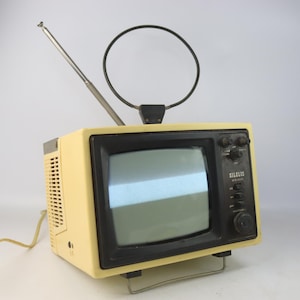 May include: A vintage cream-colored portable television with a black screen and controls. It has a telescoping antenna and a circular antenna. The brand name "SILELIS" is visible on the front. The TV sits on a metal stand.