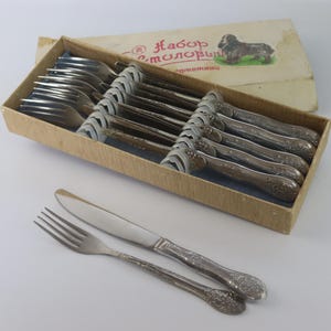May include: A vintage silverware set in a cardboard box. The set includes forks, knives, and spoons with ornate handles. The box has text and an illustration of a dog. The silverware is arranged on a blue cloth.