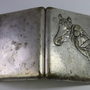 May include: A vintage silver-toned cigarette case, open to reveal a detailed relief of a horse's head and a woman's face. The case has a rectangular shape with a hinged design and shows signs of wear.