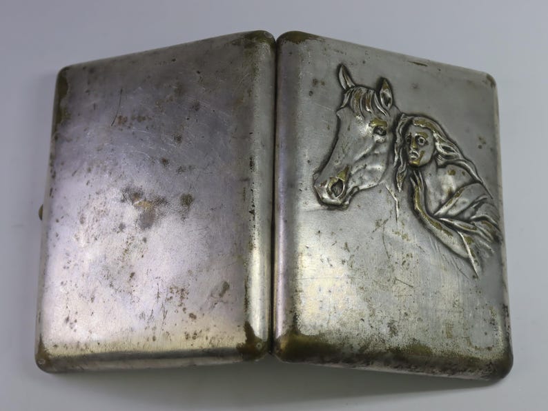 May include: A vintage silver-toned cigarette case, open to reveal a detailed relief of a horse's head and a woman's face. The case has a rectangular shape with a hinged design and shows signs of wear.