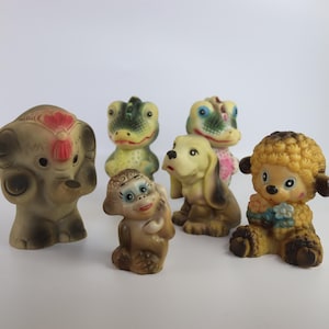 Puede incluir: Colección de juguetes de animales de goma vintage, que incluyen un elefante, un mono, un perro, una oveja y dos dinosaurios. Los juguetes son de varios tonos de marrón, beige, verde y amarillo, con detalles en rojo y azul. Los juguetes están todos de pie.