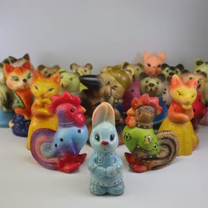 Può includere: Una collezione di figurine di animali vintage, tra cui gatti, orsi, volpi e galli. Le figurine sono dai colori vivaci, con un coniglio blu e un gallo con la cresta rossa. Questi giocattoli da collezione sono probabilmente in plastica.