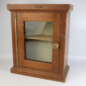 Può includere: Piccolo mobile in legno vintage con anta a vetro e un ripiano. Il mobile è in legno tinto con un dettaglio a forma di diamante sulla parte superiore. L'anta ha un piccolo pomello in legno e cerniere in ottone. Misura circa 30 cm di altezza.