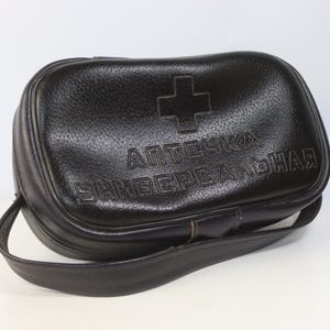 Puede incluir: Un botiquín médico rectangular de color negro con superficie texturizada, que presenta un símbolo de cruz en relieve y el texto cirílico "АПТЕЧКА УНИВЕРСАЛЬНАЯ" en la parte frontal. El bolso incluye un asa de transporte resistente en el lateral.