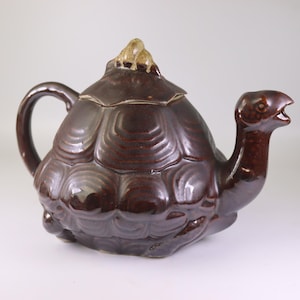 Puede incluir: Una tetera de cerámica marrón con forma de tortuga. La tetera tiene un cuerpo redondeado con un diseño de caparazón texturizado, un asa curva y un pico con forma de cabeza de tortuga. Una pequeña figura de tortuga adorna la tapa.
