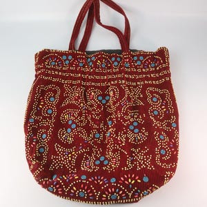Puede incluir: Un bolso tote burdeos con textura de pana. El bolso está adornado con un patrón de pequeñas cuentas de colores amarillo, azul y rosa. Tiene dos asas burdeos.