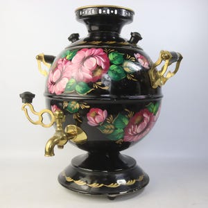 May include: A black samovar with a floral design featuring pink and red roses and green leaves. The samovar has gold-colored handles and a spigot. The base and top are also trimmed with gold accents.