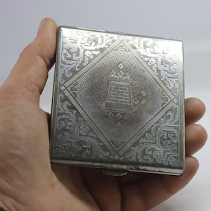 May include: A silver-toned, square compact with an ornate, engraved design. The lid features a central diamond shape with a castle motif, surrounded by a detailed geometric pattern. The compact is held in a hand.