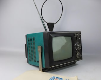 May include: A vintage teal and black portable television with a silver antenna and a circular antenna. The TV has a small screen and control knobs. A folded paper with blue and red graphics is in front of the TV.