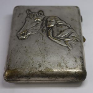 May include: A rectangular, silver-toned metal case with a hinged lid. The front features a bas-relief design of a horse's head and a woman's figure. The case has a tarnished appearance, with some areas showing a golden hue.