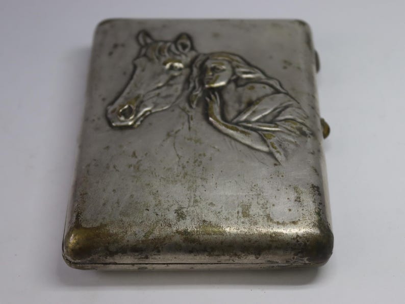 May include: A rectangular, silver-toned metal case with a hinged lid. The front features a bas-relief design of a horse's head and a woman's figure. The case has a tarnished appearance, with some areas showing a golden hue.