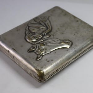 May include: A rectangular, silver-toned metal case with a detailed embossed design. The design features a stylized image of a butterfly or moth. The case has rounded corners and a slightly tarnished appearance, suggesting age.