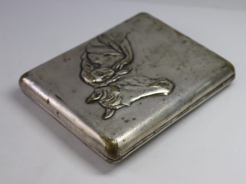May include: A rectangular, silver-toned metal case with a detailed embossed design. The design features a stylized image of a butterfly or moth. The case has rounded corners and a slightly tarnished appearance, suggesting age.