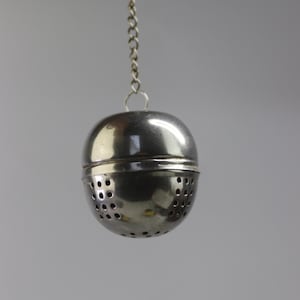 May include: A silver tea infuser, spherical in shape, suspended by a chain. The infuser is made of two halves that fit together, with small holes for steeping tea leaves. The chain is also silver and attached to the top of the infuser.